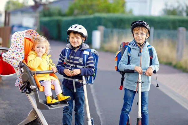 Dois miúdos da escola e uma miúda gira a ir para a escola. Pequena irmãzinha sentada no carrinho. Irmãos a andar de scooter. Feliz amilia saudável de três crianças . — Fotografia de Stock