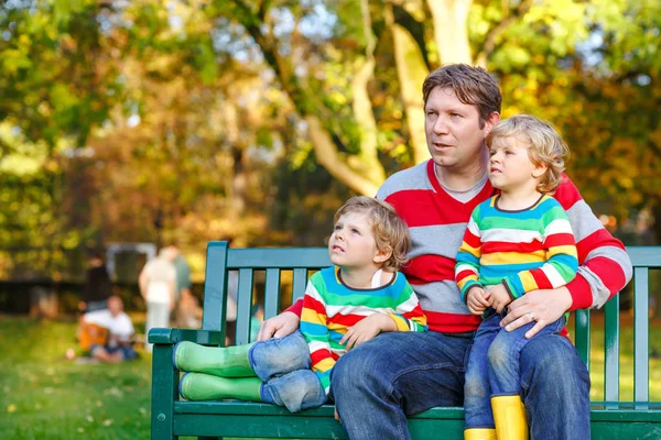 Two little kid boys and young father sitting together in colorful clothing on bench. Cute healthy children, siblings and their dad having fun in autumn park on warm sunny day. Happy family of three. — Stock Photo, Image