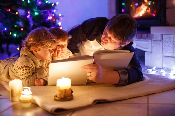 Vater und zwei kleine Jungen lesen Buch am Kamin, Kerzen und Kamin. Familie feiert Weihnachten. mit Weihnachtsbaum und Lichtern im Hintergrund. Kinder freuen sich über Bücher und Geschenke — Stockfoto