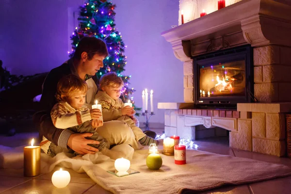 Vater und zwei kleine Jungen sitzen am Kamin, Kerzen und Kamin und schauen auf das Feuer. Familie feiert Weihnachten. Mit Weihnachtsbaum und Lichtern im Hintergrund. Kinder freuen sich über Geschenke. — Stockfoto