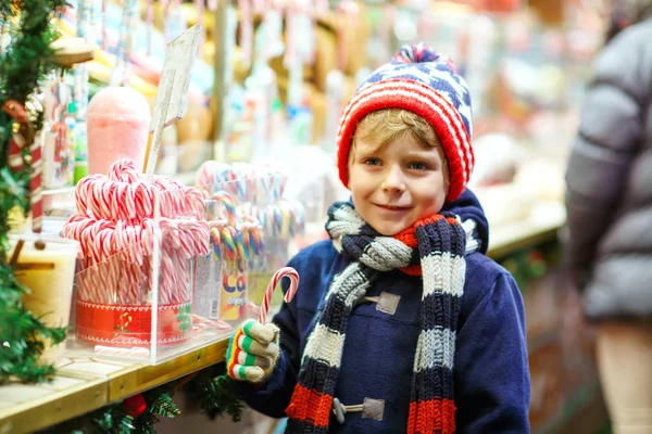 Menino bonitinho comprando doces de um stand de câncer no mercado de Natal. Criança feliz no mercado familiar tradicional na Alemanha. Pré-escolar em roupas coloridas de inverno .. — Fotografia de Stock