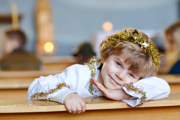 Schöner kleiner Junge, der einen Engel in der Weihnachtsgeschichte in einer Kirche spielt. glücklich liebenswert blondes Kind mit Lichtern und Weihnachtsbaum auf dem Hintergrund. Heiligabend, großer christlicher, katholischer Feiertag. — Stockfoto