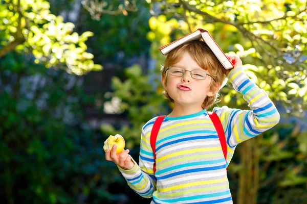Šťastný malý školáček s brýlemi, knihami, jablkem a batohem hned první den do školy nebo školky. Vtipné zdravé dítě venku za teplého slunečného dne, zpět do školy koncepce. Smějící se chlapec. — Stock fotografie
