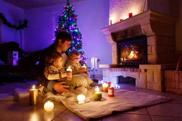 Padre e due bambini piccoli seduti accanto al camino, alle candele e al camino e che guardano il fuoco. — Foto Stock