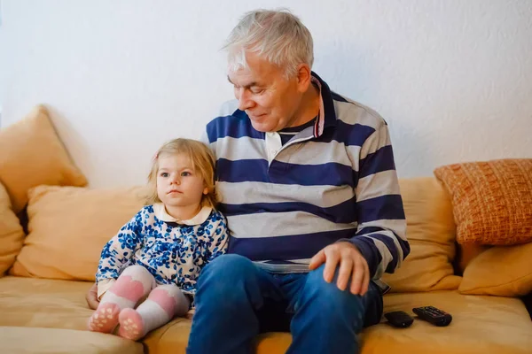 Schattig klein peuter meisje en grootvader samen kijken naar tv-show. Baby kleindochter en gelukkig voormalig senior man samen om thuis te zitten met cartoons op televisie. — Stockfoto