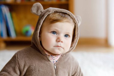 Mavi gözlü bebek, güneşli yatak odasında kahverengi ayı kışı tulumu giyiyor. Yeni doğmuş çocuk dışarıda yürümek için giyiniyor. Çocuklar için çocuk odası. Çocuklar için tekstil ve yatak. Mutlu sağlıklı kız.