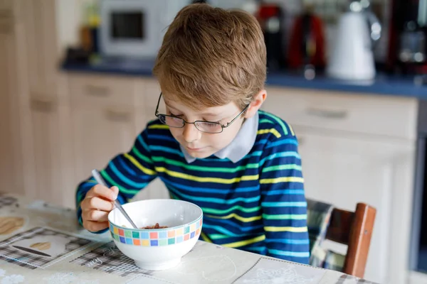 Очаровательный светловолосый мальчик в очках ест домашние хлопья на завтрак или обед. Здоровое питание для детей. В детской, в школьной столовой или дома — стоковое фото
