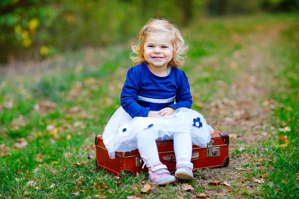 Jolie petite fille tout-petit assise sur une valise dans un parc d'automne. Joyeux bébé en bonne santé qui aime marcher avec les parents. Journée chaude ensoleillée d'automne avec enfant. Loisirs actifs et activités avec les enfants dans la nature . — Photo