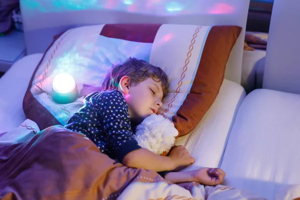 A kis óvodás fiú színes lámpával alszik az ágyban. Iskolás gyerek álmodik, és kezében plüss játék. A gyerek dühös a sötétségre — Stock Fotó