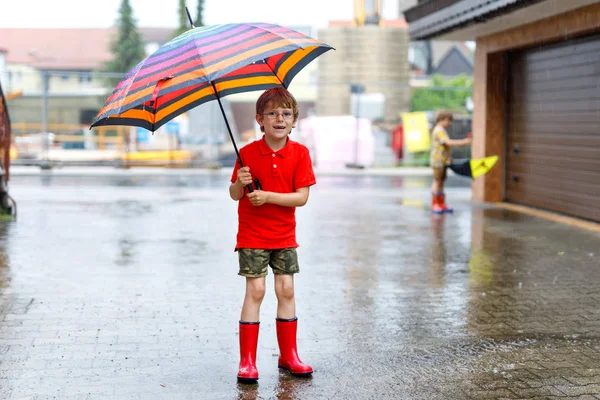 Junge in roten Regenstiefeln und mit buntem Regenschirm auf der Straße der Stadt. Kind mit Brille an einem Sommertag. glückliches Kind bei starkem Sommerregen. — Stockfoto