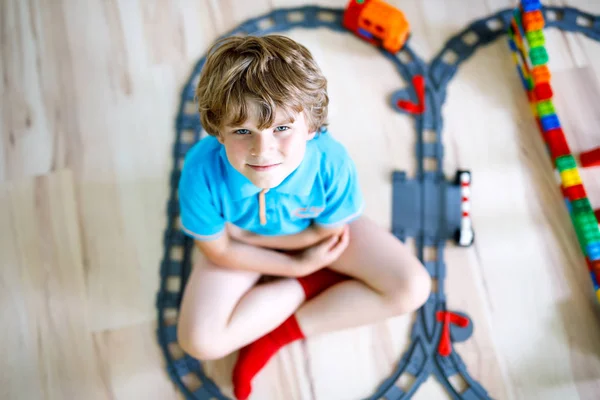 Entzückender kleiner blonder Junge, der mit bunten Plastikklötzen spielt und einen Bahnhof erschafft. Kind hat Spaß beim Bau von Eisenbahnspielzeug zu Hause.. — Stockfoto