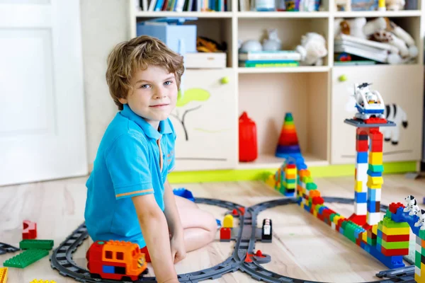 Bedårande liten blond unge pojke leker med färgglada plast block och skapa tågstation. Barn ha roligt med att bygga järnväg leksaker hemma.. — Stockfoto