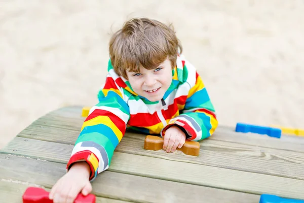 Niño rubio feliz divirtiéndose y escalando en el patio al aire libre. Divertido niño alegre sonriendo y haciendo deportes. Vacaciones de verano, primavera y otoño para niños activos. Niño en ropa de moda colorida . — Foto de Stock