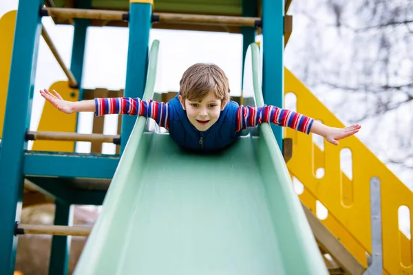 Happy blond kid boy having fun and sliding on outdoor playground. Funny joyful child smiling and climbing on slide. Summer, spring and autumn leisure for active kids. Boy in colorful casual clothes. — Stock Photo, Image