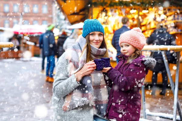 Menina criança feliz e jovem mulher bonita com xícara de chocolate quente fumegante e vinho quente. Criança adorável e mãe bonita no mercado de Natal na Alemanha. Família caminhando no mercado de Natal . — Fotografia de Stock