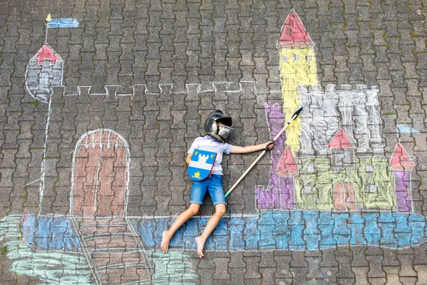 Little active kid boy drawing knight castle and fortress with colorful chalks on asphalt. Happy child in helmet and with spear having fun with playing knight game and painting — Stock Photo, Image