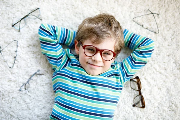 Retrato de cerca del pequeño niño rubio con diferentes anteojos sobre fondo blanco. Feliz niño sonriente con ropa casual. Infancia, visión, gafas, tienda de óptica. Niño elegir nuevas gafas . —  Fotos de Stock