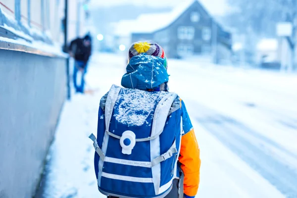 Kleine schooljongen van de lagere klasse die naar school loopt tijdens de sneeuwval. Vroege ochtend en besneeuwde straten in de stad. Leerling met rugzak of tas in kleurrijke winterkleding. — Stockfoto