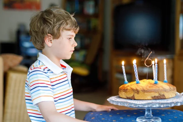 Entzückend glücklicher blonder kleiner Junge, der seinen Geburtstag feiert. Kind bläst Kerzen auf selbstgebackenem Kuchen, drinnen. Geburtstagsfeier für Schulkinder, Familienfeier ab 6 Jahren — Stockfoto