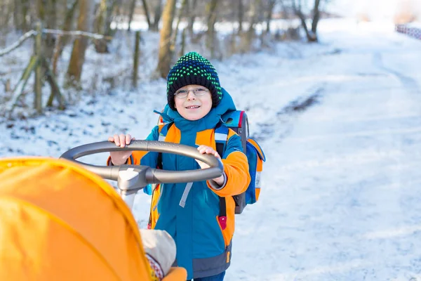 Proud little kid boy pushing stroller with newborn baby. Happy sibling and brother, on the way from school on sunny winter day. Child with colorful clothes and orange pram and backpack school stuff — Stock Photo, Image