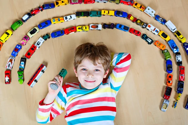 Aranyos kis szőke fiú, aki sok játékautóval játszik idebent. A gyerek színes inget visel. Boldog óvodás, aki otthon vagy óvodában szórakozik. Különböző járművek nagy gyűjteménye. Boldogság játék. — Stock Fotó