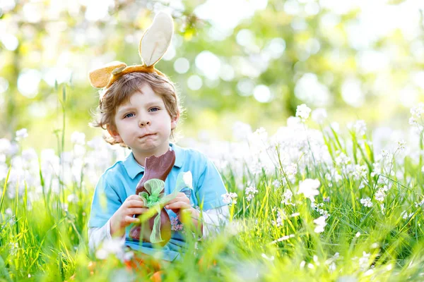 Cute little kid boy with Easter bunny ears celebrating traditional feast. Happy child eating chocolate rabbit fugure on warm sunny day. Family, holiday, spring concept. Toddler sitting between flowers — Stock Photo, Image