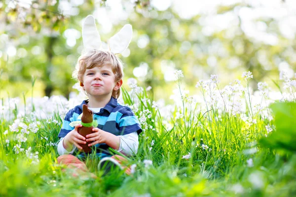 Cute little kid boy with Easter bunny ears celebrating traditional feast. Happy child eating chocolate rabbit fugure on warm sunny day. Family, holiday, spring concept. Toddler sitting between flowers — Stock Photo, Image