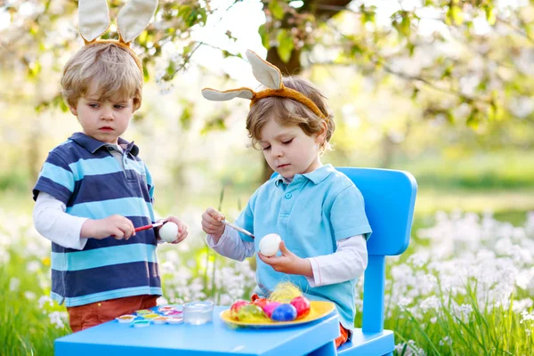 Два милых маленьких мальчика с пасхальными кроличьими ушами, красили красочные яйца и веселились на открытом воздухе. Семья, братья, братья, близнецы празднуют религиозный праздник. Лучшие друзья раскрашивают яйца. — стоковое фото