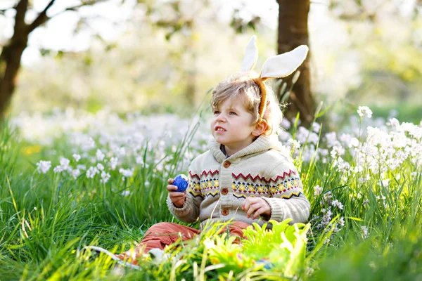 Słodki chłopczyk z króliczymi uszami bawiący się tradycyjnymi polowaniami na pisanki w ciepły słoneczny dzień, na świeżym powietrzu. Świętujemy Wielkanoc. Znalezienie malucha, kolorowe jaja w zielonej trawie — Zdjęcie stockowe