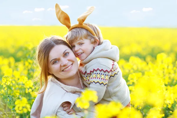 Pieni lapsi ja hänen äitinsä pääsiäispupun korvissa hauskaa, juhlivat perinteistä pääsiäislomaa. Lapsipoika ja äiti, nainen keltaisella raiskauskentällä, ulkona. Perhe yhdessä, rakkaus, onnellisuus. — kuvapankkivalokuva