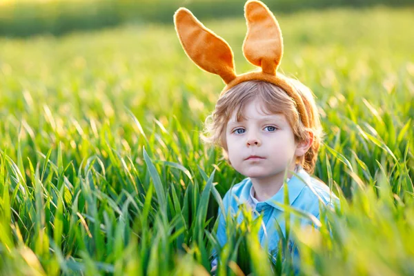 Cute little kid boy with bunny ears having fun with traditional Easter eggs hunt on warm sunny day, outdoors. Celebrating Easter holiday. Toddler finding, colorful eggs in green grass.. — Stock Photo, Image