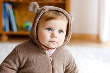 Mavi gözlü bebek, güneşli yatak odasında kahverengi ayı kışı tulumu giyiyor. Yeni doğmuş çocuk dışarıda yürümek için giyiniyor. Çocuklar için çocuk odası. Çocuklar için tekstil ve yatak. Mutlu sağlıklı kız.
