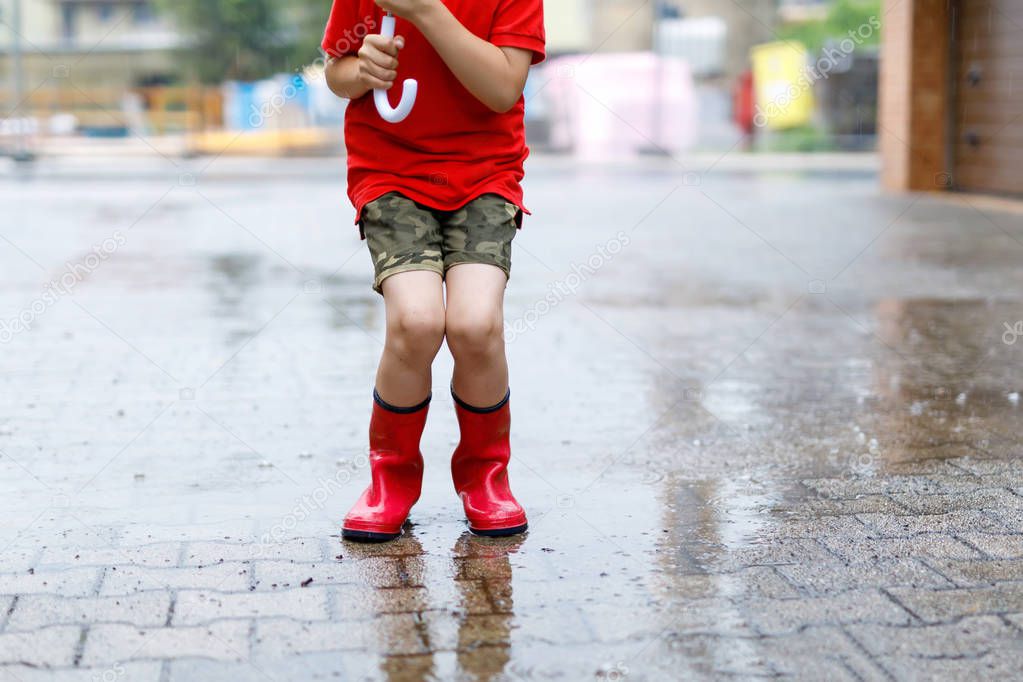 Child wearing red rain boots jumping into a puddle. Close up. Kid having fun with splashing with water. Warm heavy summer rain and happy children.