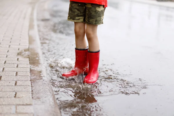Su birikintisine atlayan kırmızı çizme giyen bir çocuk. Kapatın. Çocuk su sıçratarak eğleniyor. Sıcak yaz yağmuru ve mutlu çocuklar.