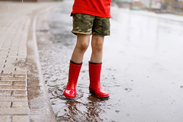 Criança usando botas de chuva vermelha pulando em uma poça. Fecha. Miúdo a divertir-se com salpicos de água. Chuva quente de verão pesado e crianças felizes. — Fotografia de Stock