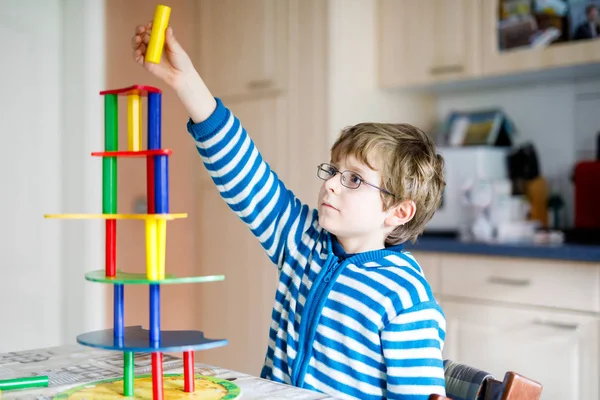 Blond child with glasses playing with lots of colorful wooden blocks game indoor. Active funny kid boy having fun with building and creating, balance toy. Home, kidsroom. Cognitive development.