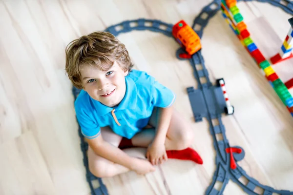 Bedårande liten blond unge pojke leker med färgglada plast block och skapa tågstation. Barn ha roligt med att bygga järnväg leksaker hemma.. — Stockfoto