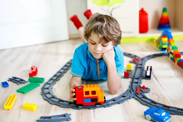 Adorabile ragazzino biondo che gioca con blocchi di plastica colorati e crea la stazione ferroviaria. Bambino divertirsi con la costruzione di giocattoli ferroviari a casa .. — Foto Stock