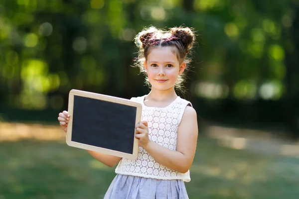 Glad liten flicka som håller tomma krita skrivbord i händerna. Skolbarn på första dagen av grundskolan. Friska bedårande barn utomhus, i grön park. copyspace på skrivbordet — Stockfoto