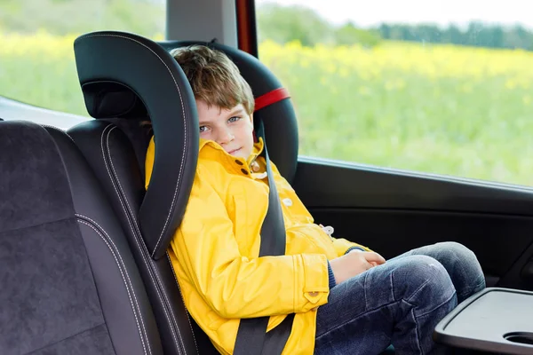 Adorable cute preschool kid boy sitting in car in yellow rain coat. Little school child in safety car seat with belt enjoying trip and jorney. Safe travel with kids and traffic laws concept — Stock Photo, Image