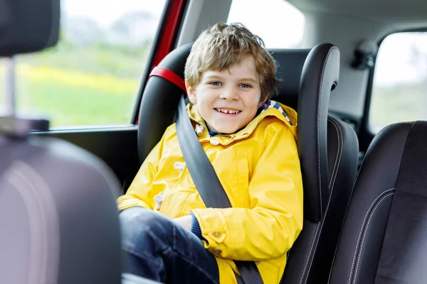 Roztomilý roztomilý předškolní chlapec sedí v autě ve žlutém plášti. Malá školačka v autosedačce s pásem si užívá výlet a výlet. Bezpečné cestování s dětmi a dopravní zákony koncept — Stock fotografie