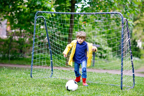 Actieve schattige kleine jongen jongen spelen voetbal en voetbal en plezier, buitenshuis. Kind spel op regenachtige dag. Het dragen van geel regen jas en rood rubber laarzen. — Stockfoto