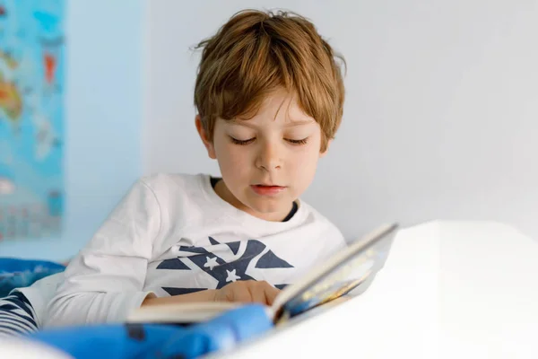 Lindo niño rubio en pijama leyendo un libro en su dormitorio. Un niño emocionado leyendo alto, sentado en su cama. Colegial, familia, educación —  Fotos de Stock