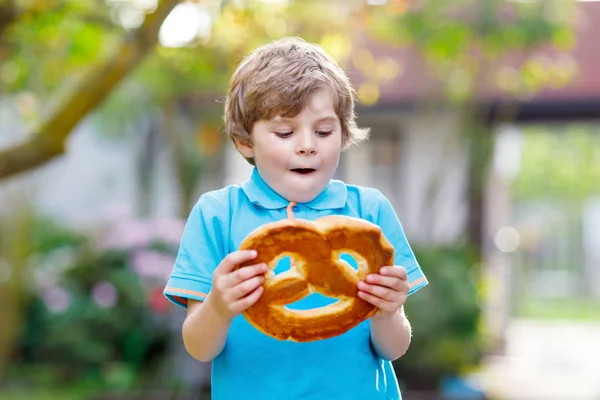 En förtjusande liten pojke som äter en stor bayersk kringla. Glad blond barn njuter av smakfulla traditionella bröd. Hälsosam mat för glada barn — Stockfoto