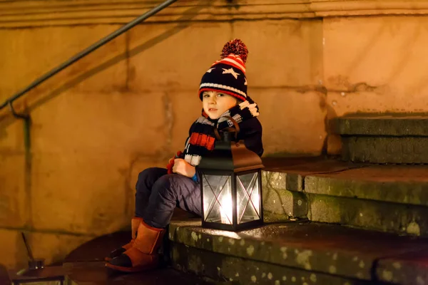 Pequeno menino bonito com uma lanterna de luz nas escadas perto da igreja. Criança feliz no mercado de Natal na Alemanha. Criança à espera dos pais no dia frio de inverno — Fotografia de Stock