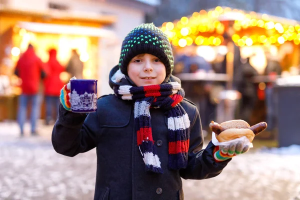 Little cute kid boy eating German sausage and drinking hot children punch on Christmas market. Happy child on traditional family market in Germany, Munich. Laughing boy in colorful winter clothes — Stock Photo, Image
