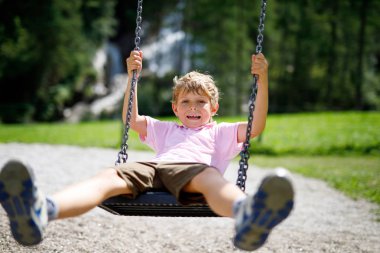 Komik çocuk, zincire vurulup ıslanırken dışarıda oyun alanında sallanırken eğleniyor. Çocuk yaz günü sallanıyor. Çocuklarla aktif zaman geçirme. Yüzüne yağmur damlaları damlayan mutlu ağlayan çocuk.