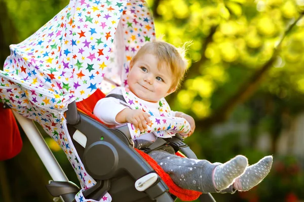 Linda niña hermosa y saludable sentada en el cochecito o cochecito y esperando a mamá. Feliz niño sonriente con ojos azules. Con fondo de árbol verde. Hija pequeña va a dar un paseo con la familia — Foto de Stock