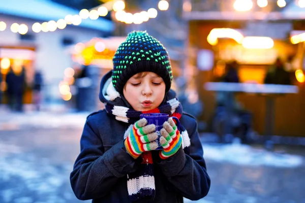 Kleiner süßer Junge trinkt heißen Kinderpunsch oder Schokolade auf dem deutschen Weihnachtsmarkt. Glückliches Kind auf traditionellem Familienmarkt in Deutschland, Lachender Junge in bunter Winterkleidung — Stockfoto