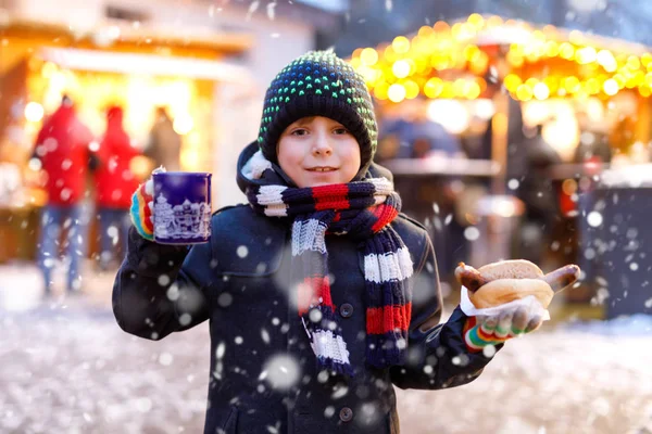 Menino bonito comendo salsicha alemã e bebendo soco crianças quentes no mercado de Natal. Criança feliz no mercado familiar tradicional na Alemanha, Munique. Garoto rindo em roupas coloridas de inverno — Fotografia de Stock